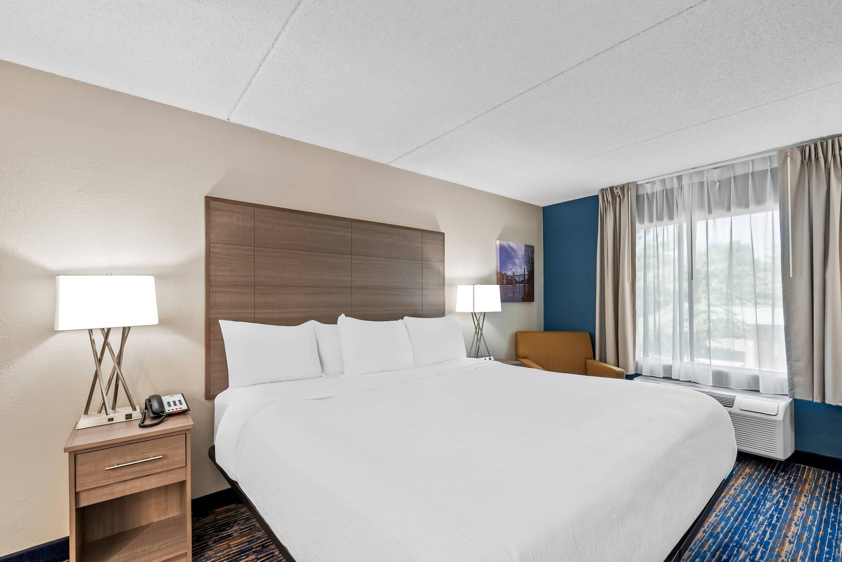 A hotel room with a large bed, two bedside tables with lamps, a chair, a window with curtains, and a wall-mounted air conditioner.