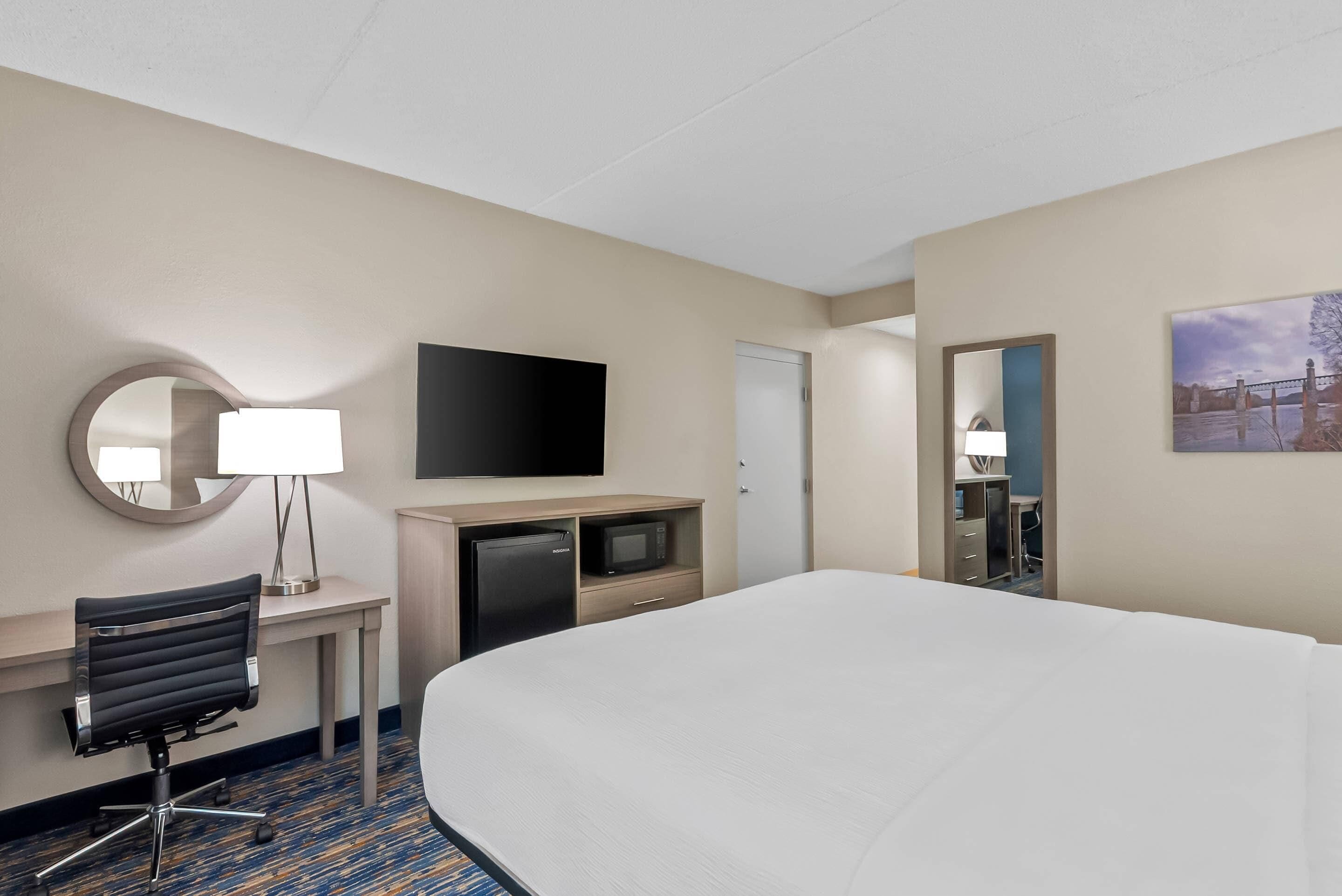 Quarto de hotel com cama, escrivaninha, cadeira, televisão e espelho.