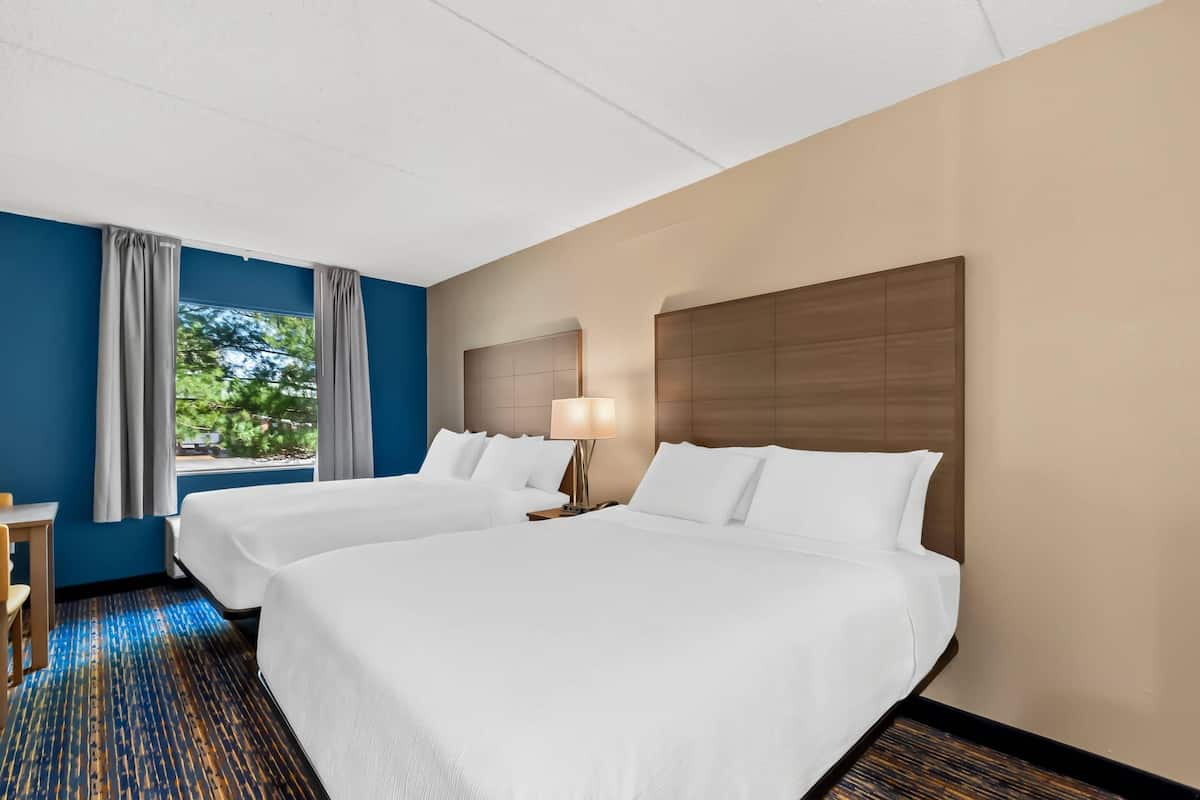 A hotel room with two beds, a desk, a lamp, and a view of greenery.