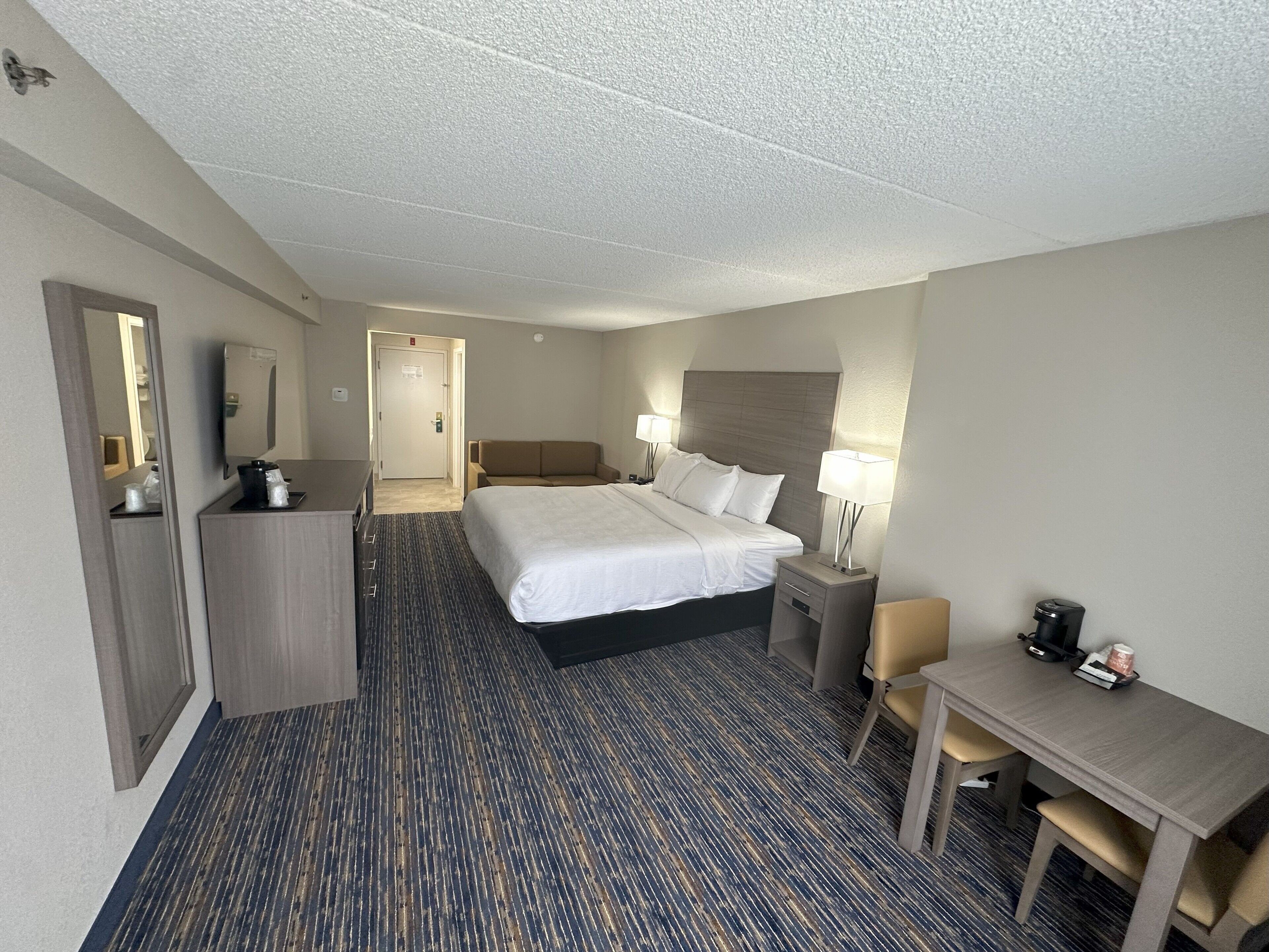 A hotel room with a large bed, a desk, a chair, a dresser, and a mirror.