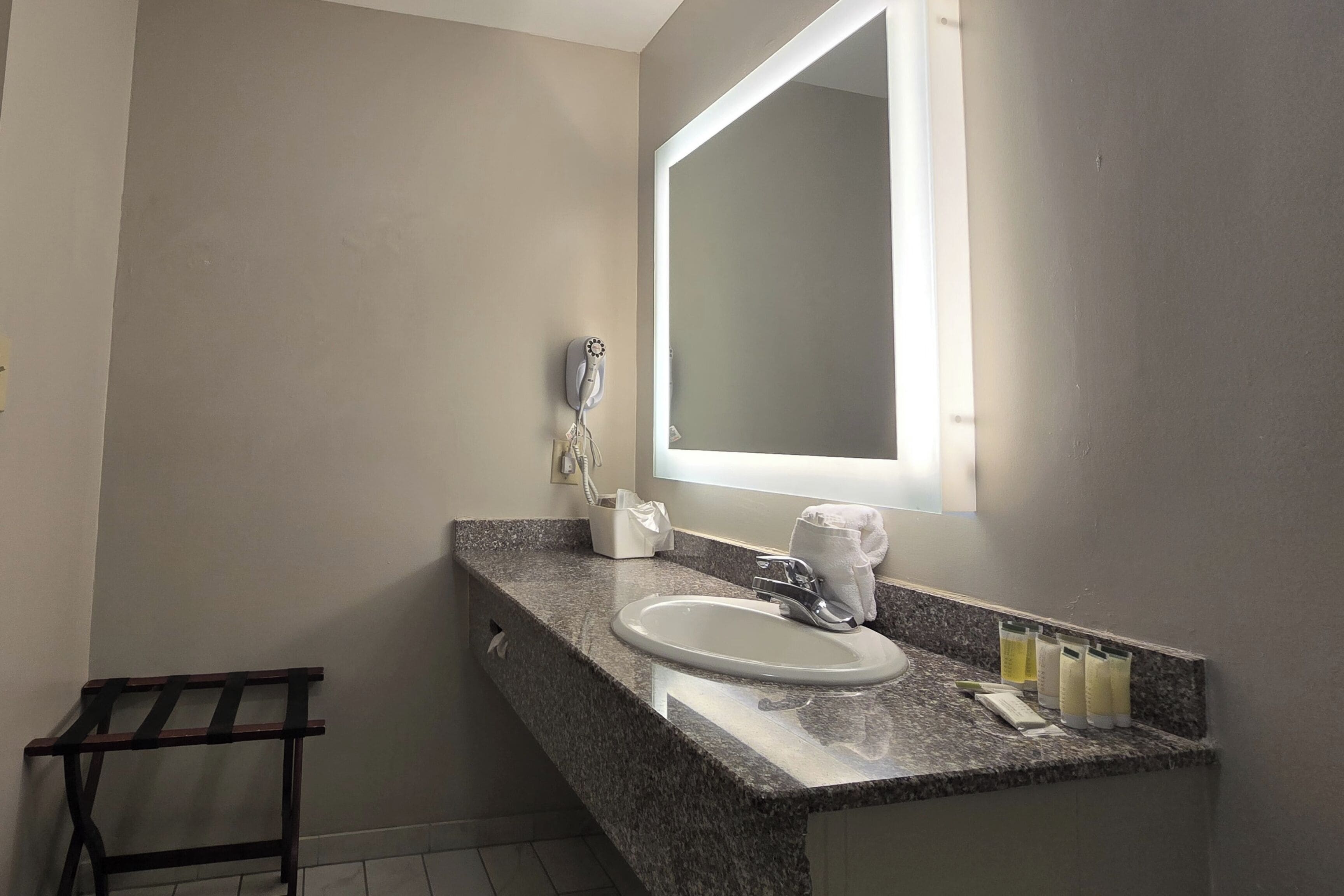 A bathroom with a granite countertop, a white sink, a mirror, a hairdryer, and toiletries.