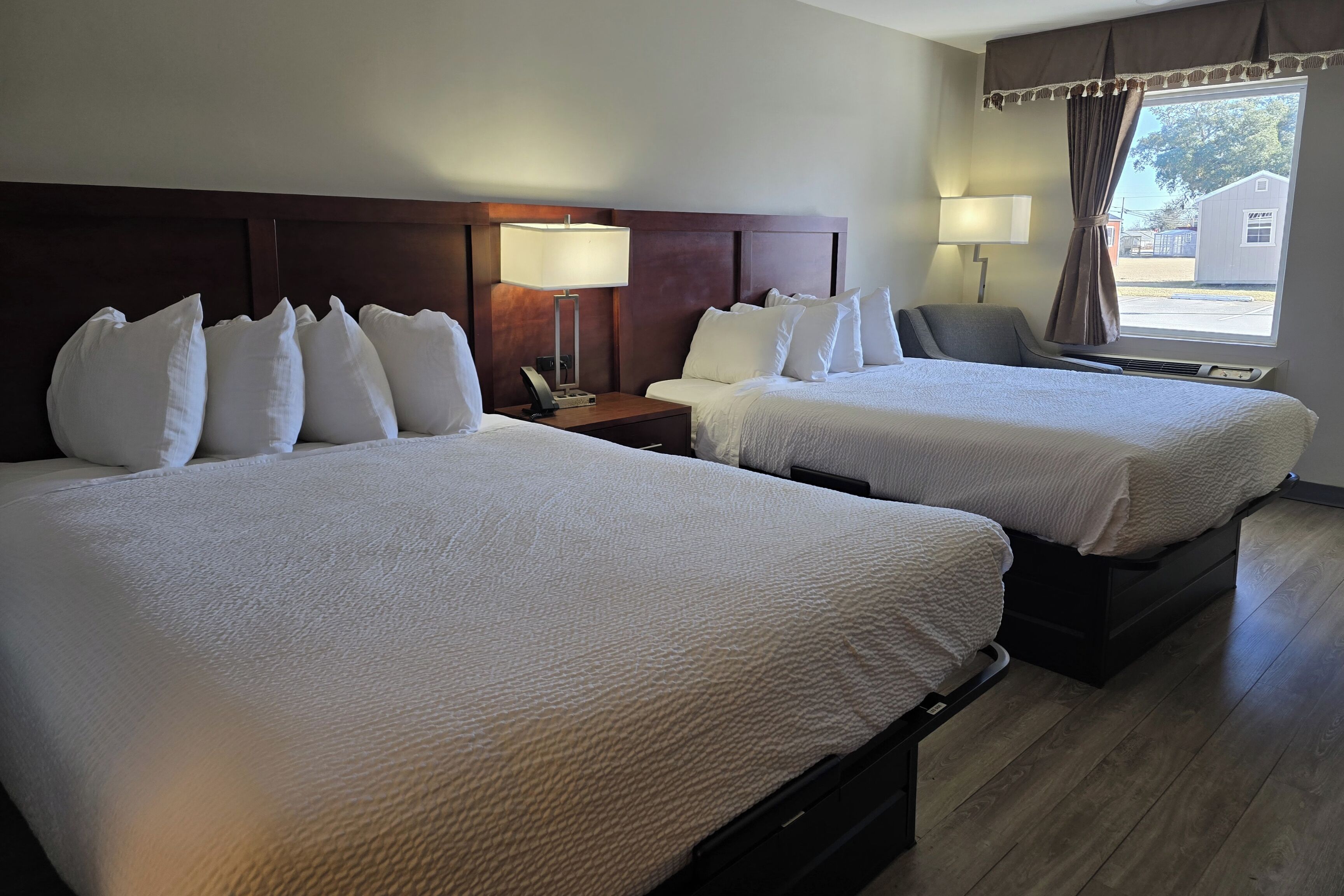 A hotel room with two beds, a nightstand with a lamp, a chair, and a window with curtains.