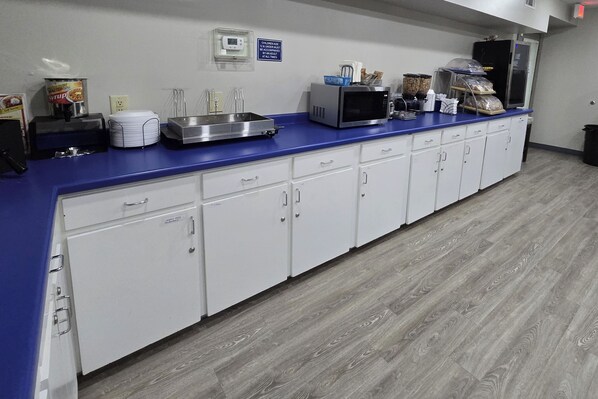 Uma área de cozinha com bancada azul, armários brancos, micro-ondas, máquina de café e um letreiro na parede.