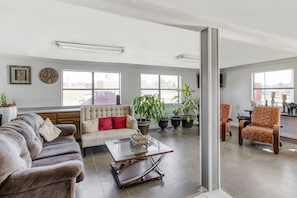 Uma sala de estar ampla com sofá, poltronas e mesa de centro. Há janelas grandes e plantas em vasos.