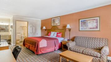 A hotel room with a bed, a sofa, a desk, and a chair. There is a bathroom with a toilet and a sink.