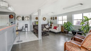 A spacious living area with a sofa, armchairs, and a coffee table. There are several potted plants and a large window providing natural light.