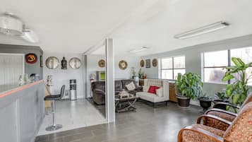 A spacious living area with a sofa, armchairs, and a coffee table. There are several potted plants and a large window providing natural light.