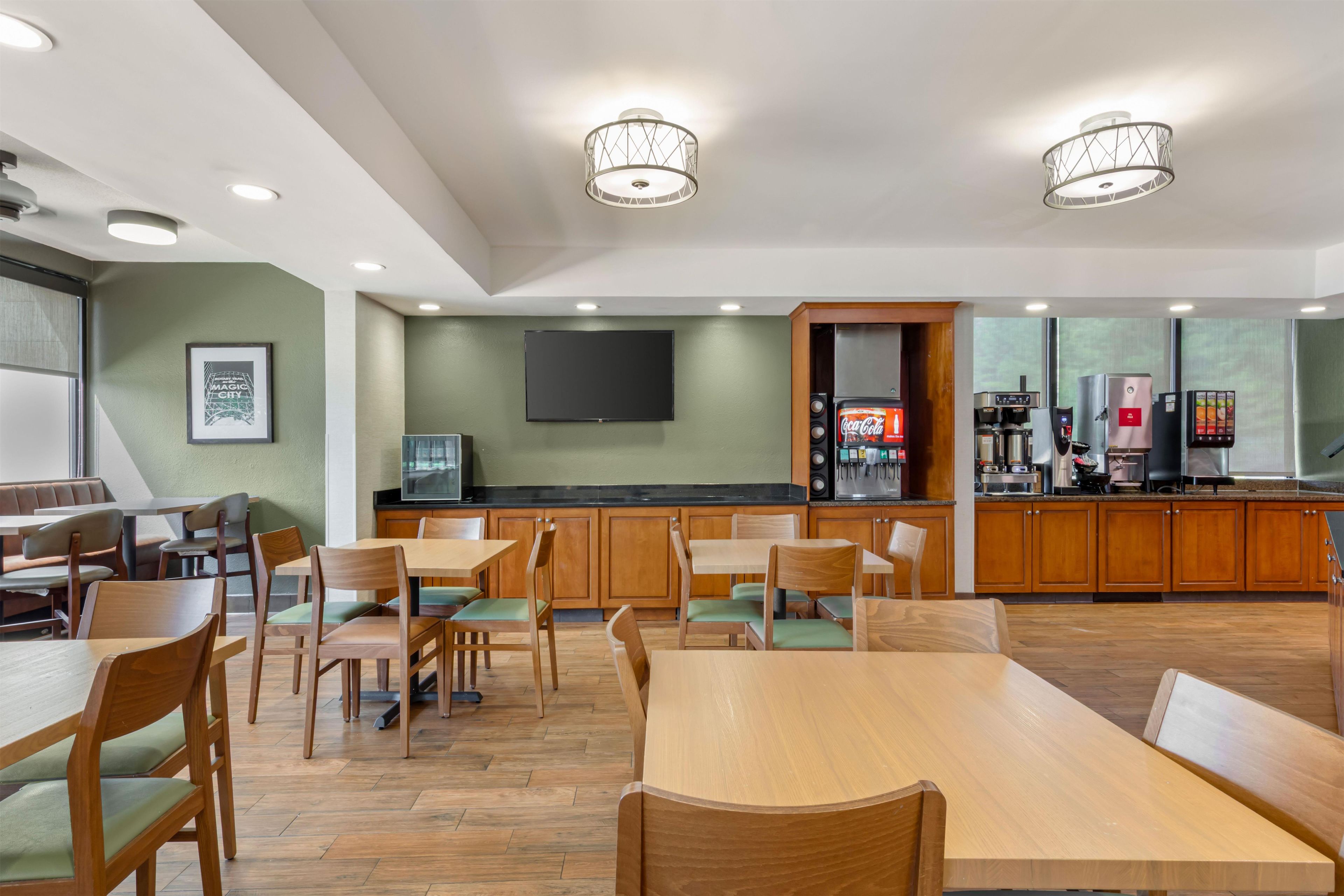 A modern dining area with wooden tables and chairs, a flat-screen TV mounted on the wall, and a coffee machine in the background.