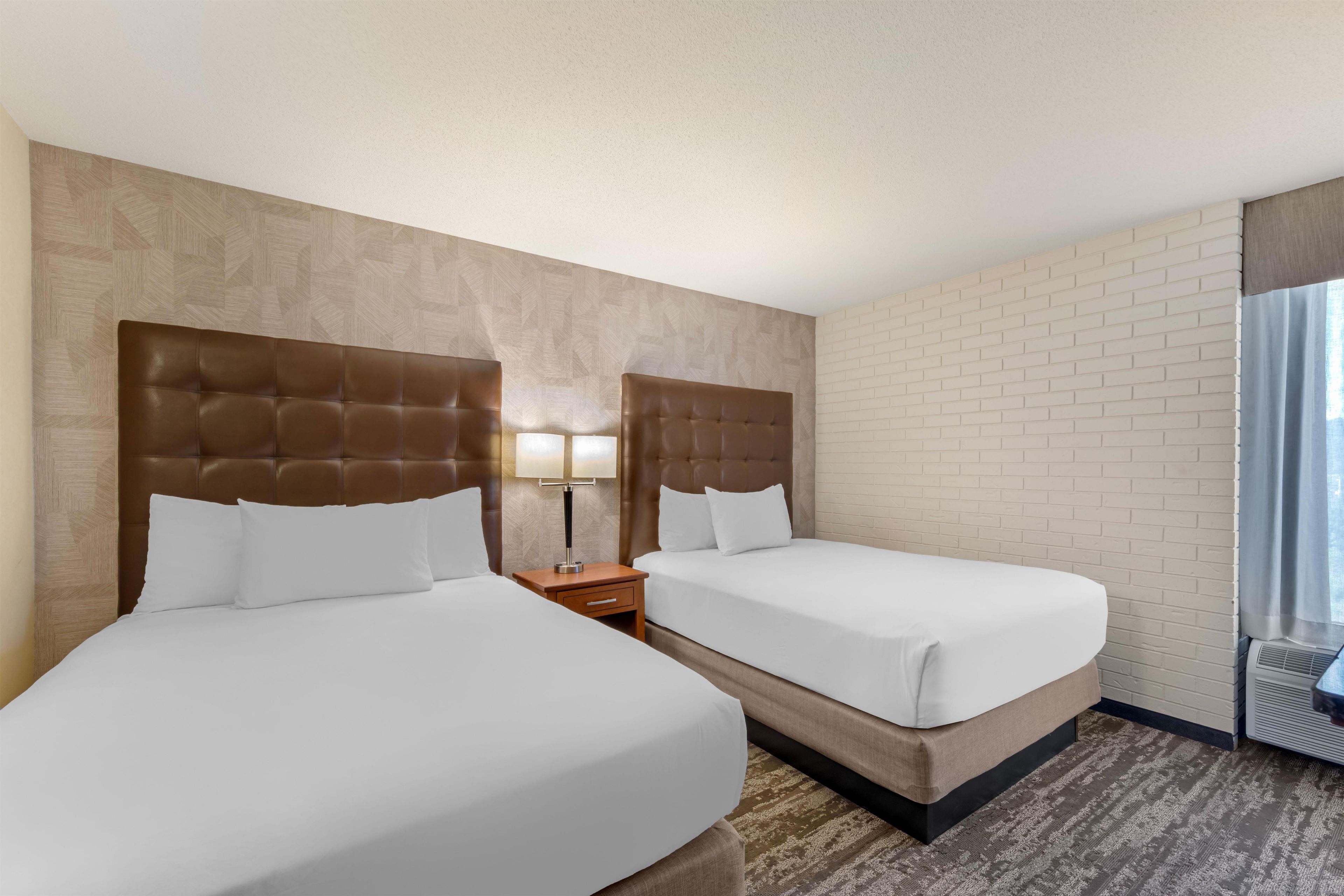 A hotel room with two beds, a nightstand with a lamp, a brick-patterned wall, and a window with curtains.