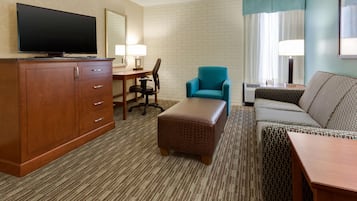 A hotel room with a flat-screen TV on a wooden cabinet, a desk with a chair, a teal armchair, a patterned sofa, and a small wooden table.