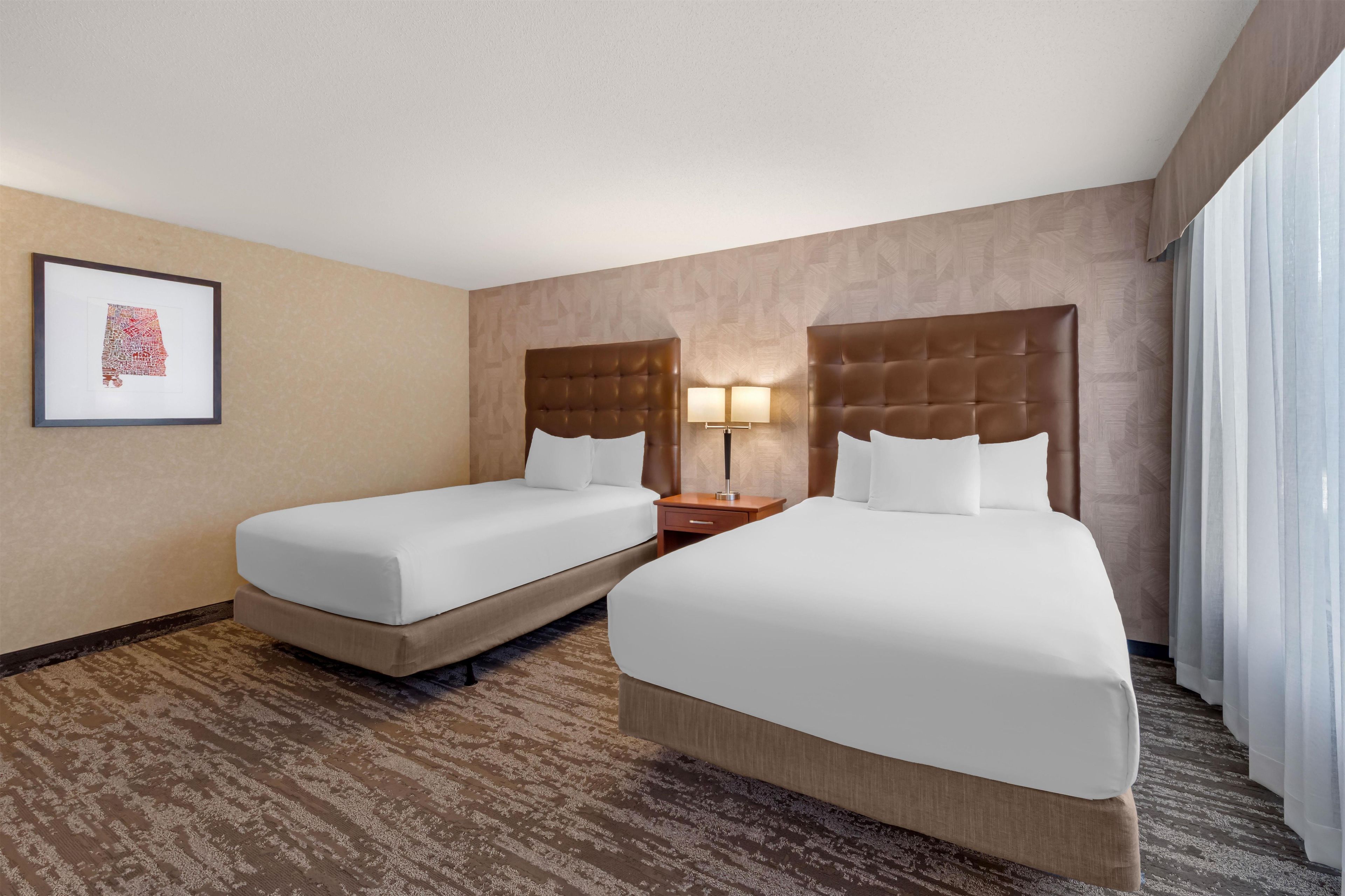A hotel room with two beds, a nightstand with a lamp, a framed picture on the wall, and a patterned carpet.