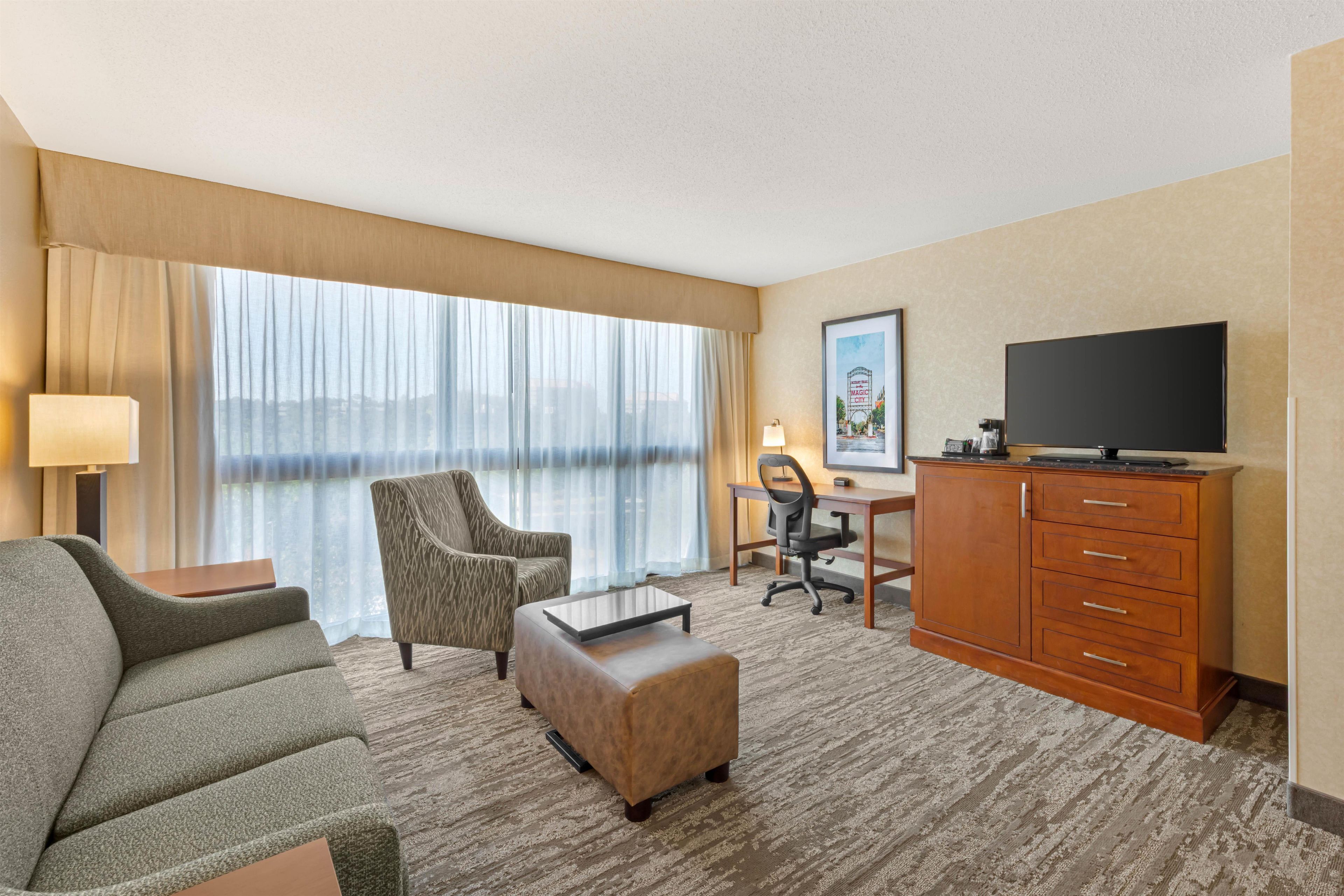 A hotel room with a sofa, armchair, desk, chair, ottoman, television, and a large window with curtains.