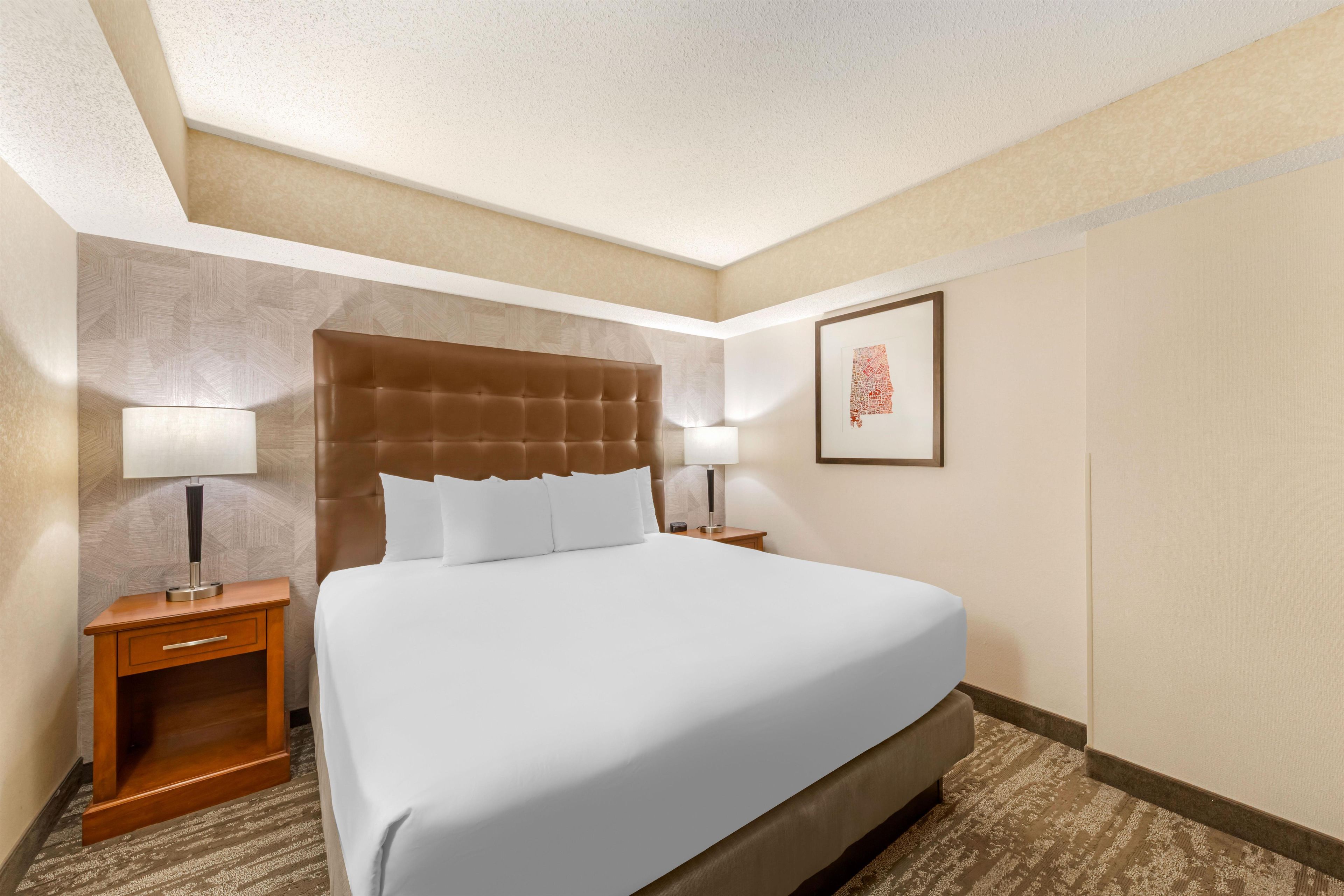 A hotel room with a large bed, two bedside tables with lamps, a wooden nightstand, and a framed picture on the wall.
