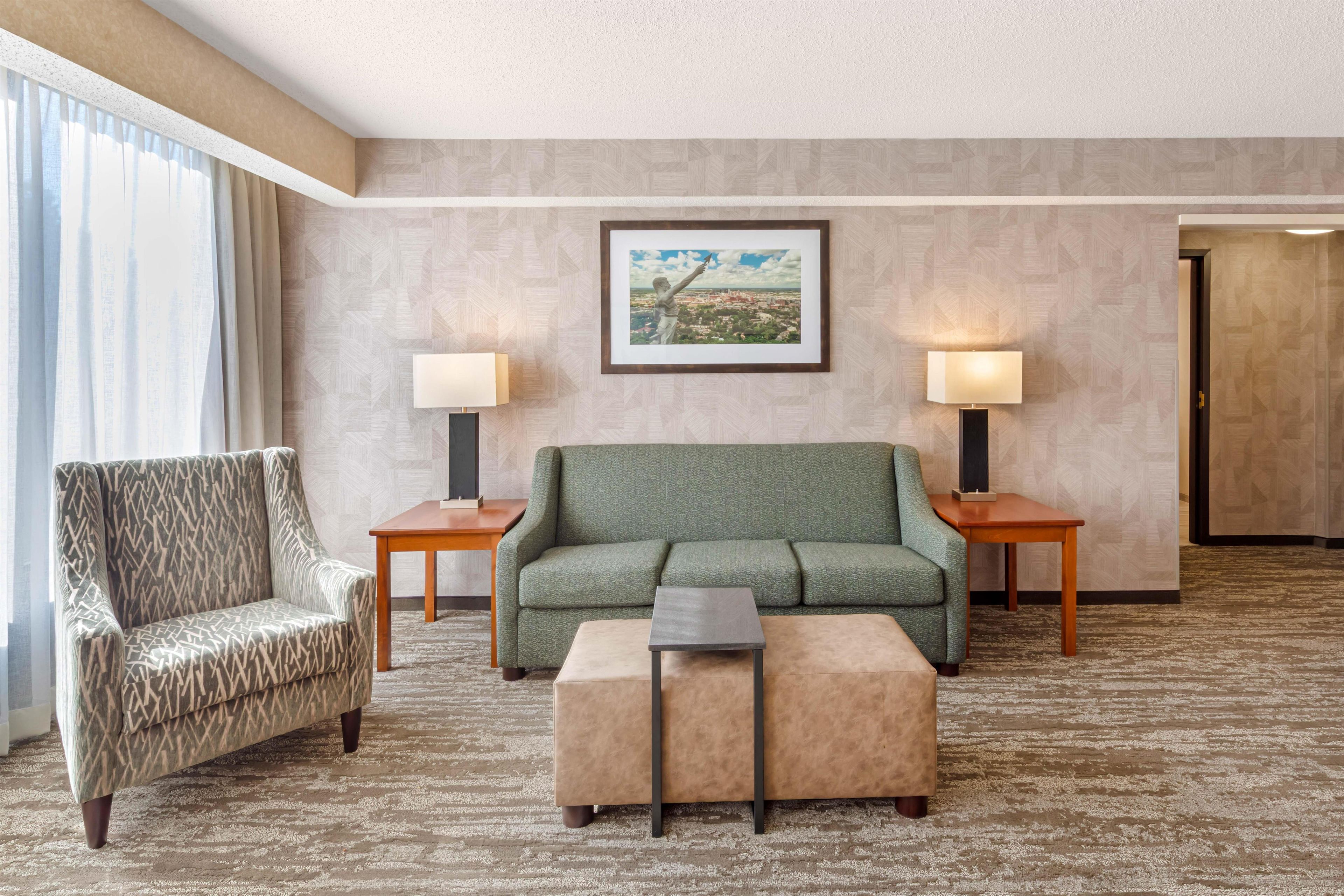 Habitación de hotel con un sillón estampado, un sofá verde, un pequeño reposapiés, dos lámparas de noche y una fotografía enmarcada de un paisaje urbano en la pared.