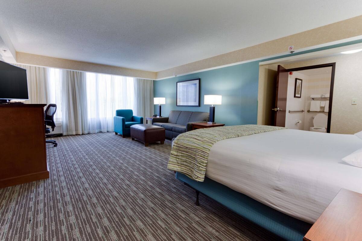 A hotel room with a large bed, a desk with a television, a sofa, a chair, and a bathroom visible through an open door.