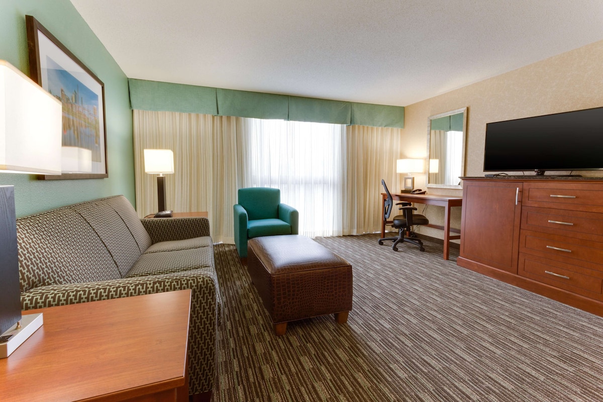 A hotel room with a sofa, a teal armchair, a wooden desk, a television, and a large window with curtains.