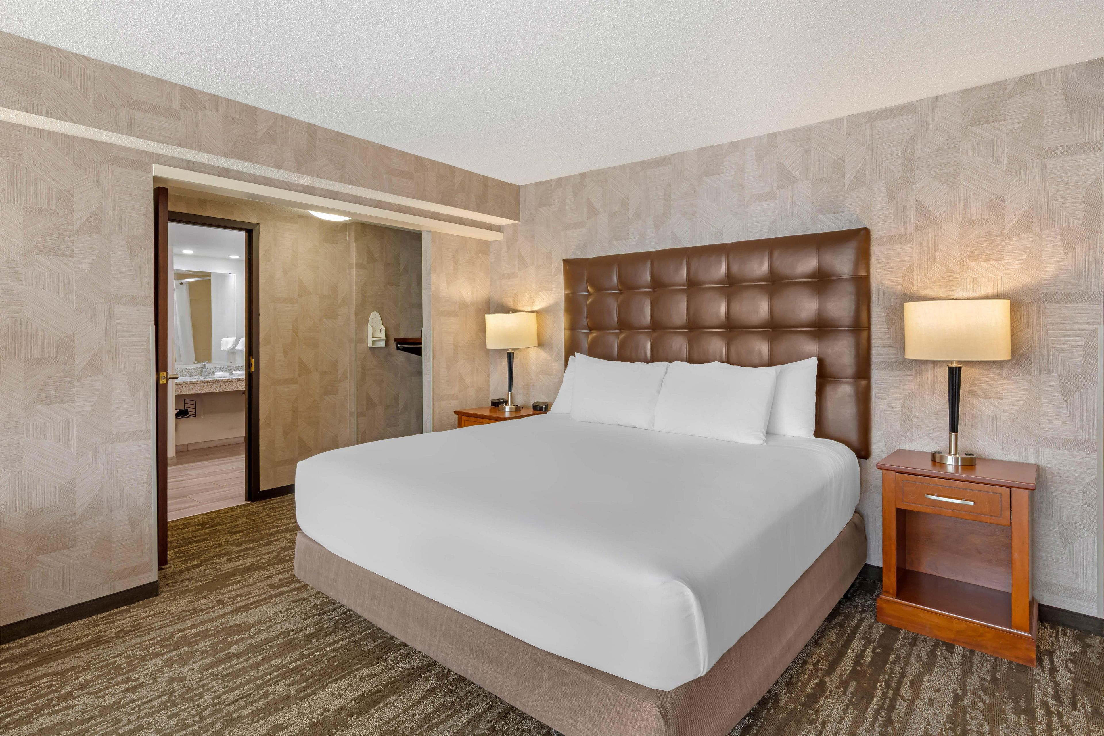A hotel room with a large bed, two bedside tables with lamps, a wooden nightstand, a mirror, and a door leading to a bathroom.