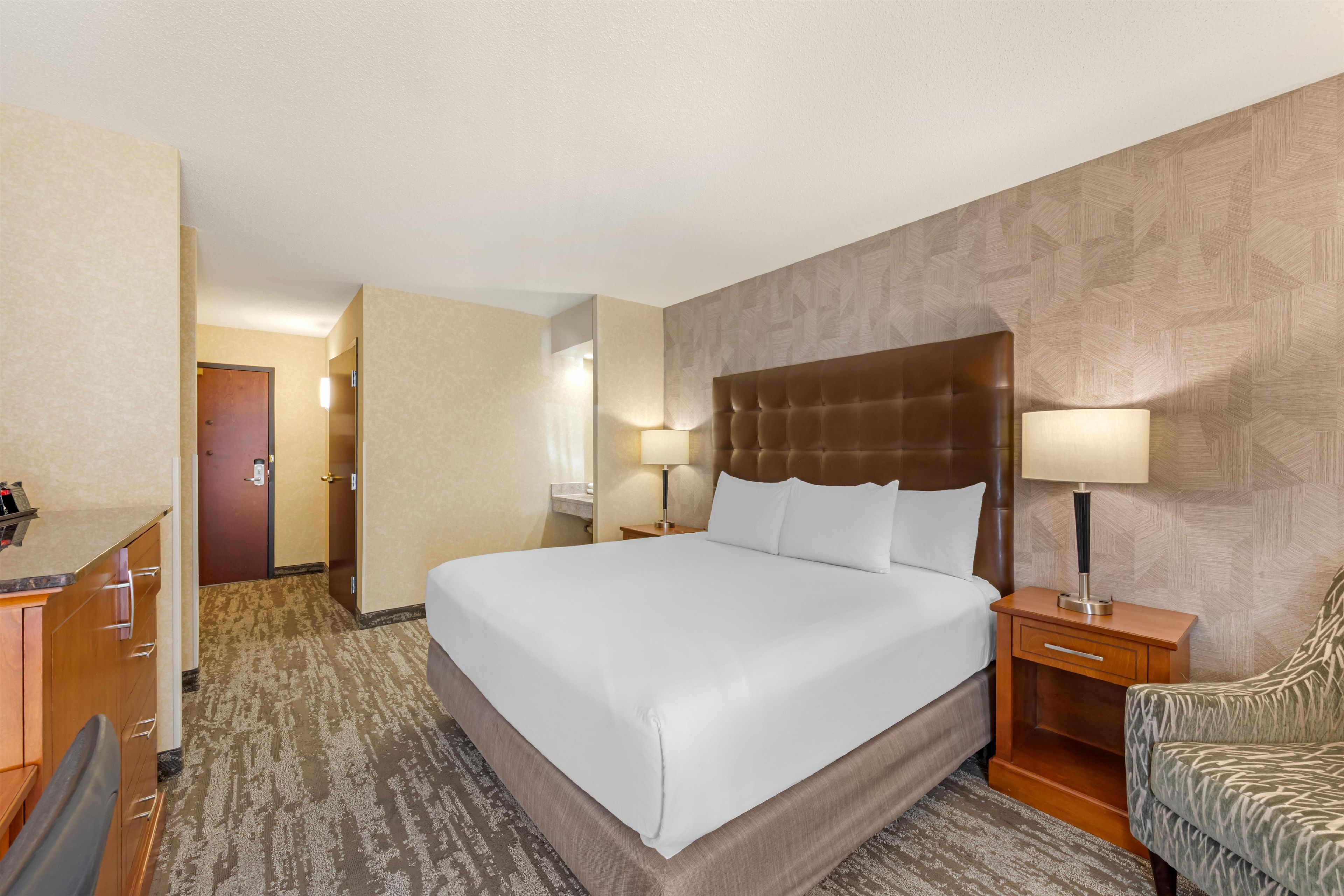 A hotel room with a large bed, a desk, a chair, a nightstand, and a bathroom visible through an open door.