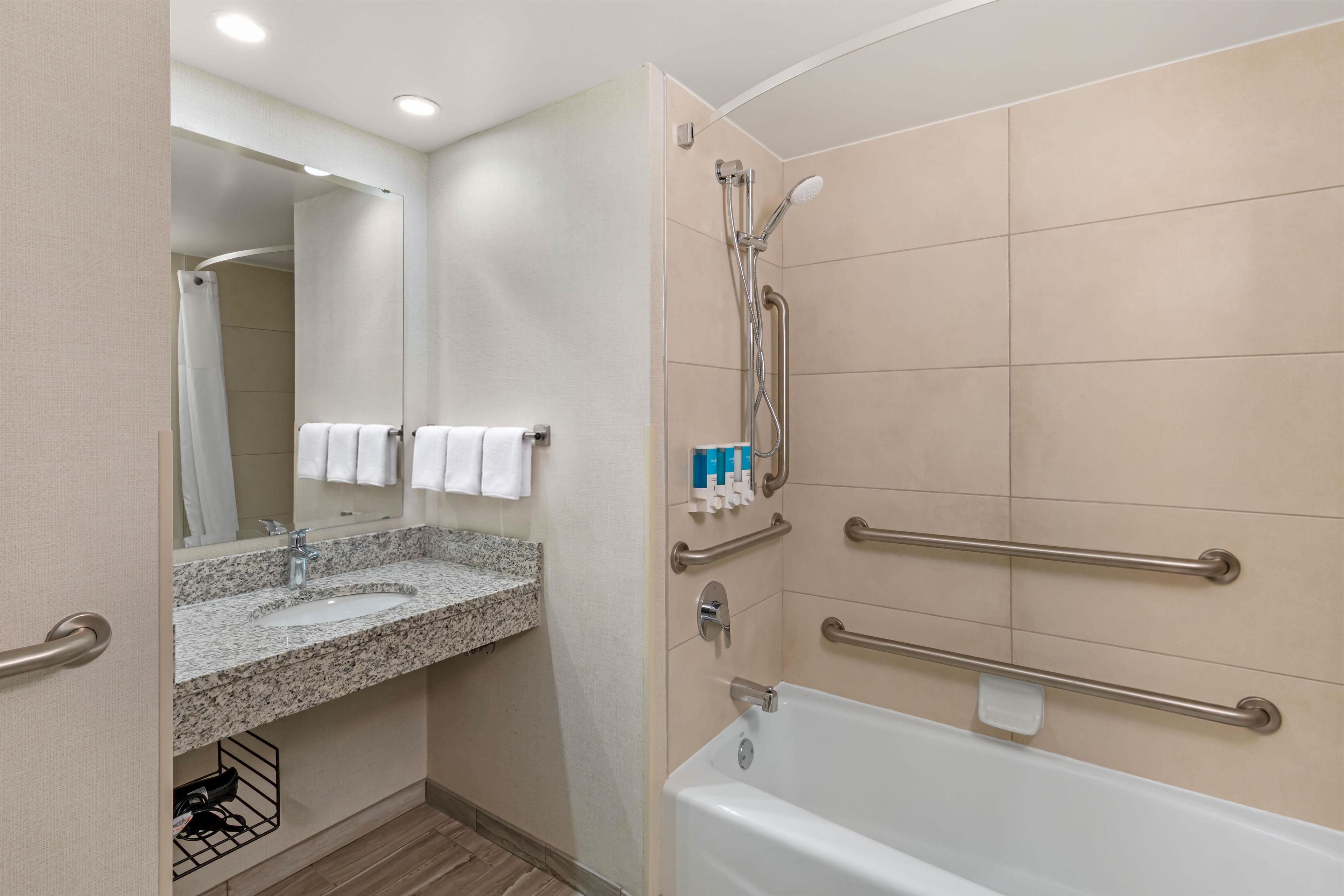 A bathroom with a granite sink, a mirror, a shower with grab bars, and a white bathtub.