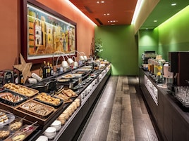 Disposição de buffet com diversos alimentos e bebidas, um quadro na parede e uma parede verde decorativa.