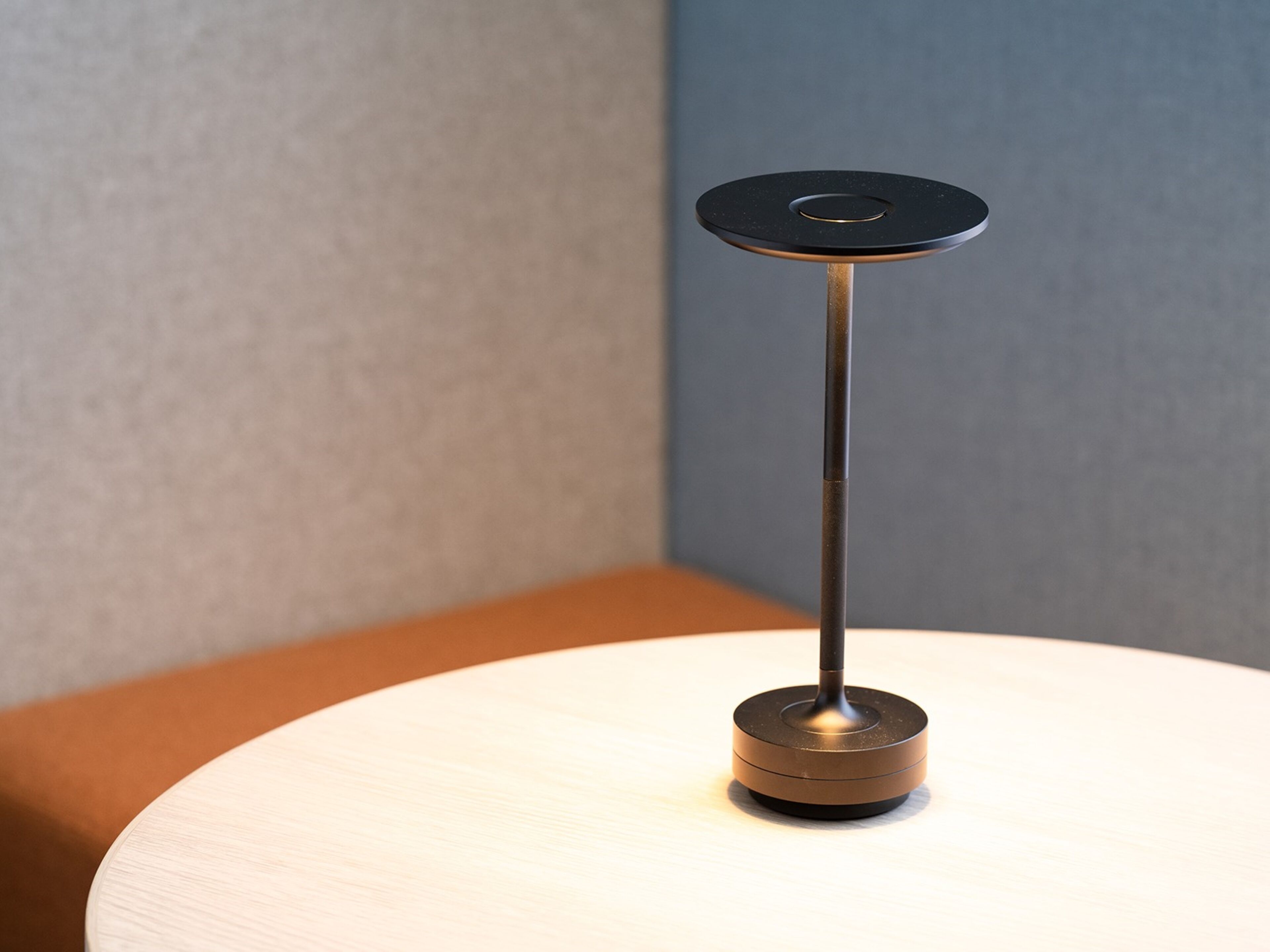 A modern table lamp with a metallic base and a black circular shade, placed on a round table against a neutral-colored wall.