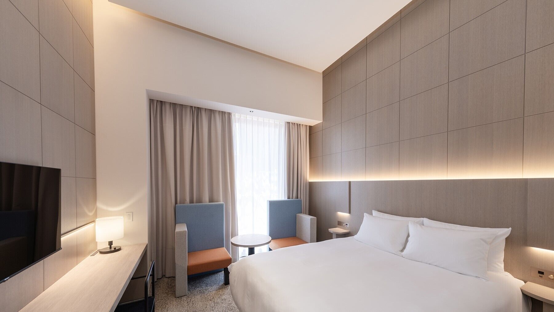 A modern hotel room with a large bed, a desk, a chair, a small table, and a TV.