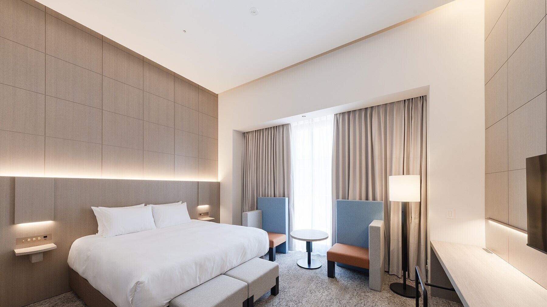 A modern hotel room with a large bed, a desk, a chair, a small table, and a television.