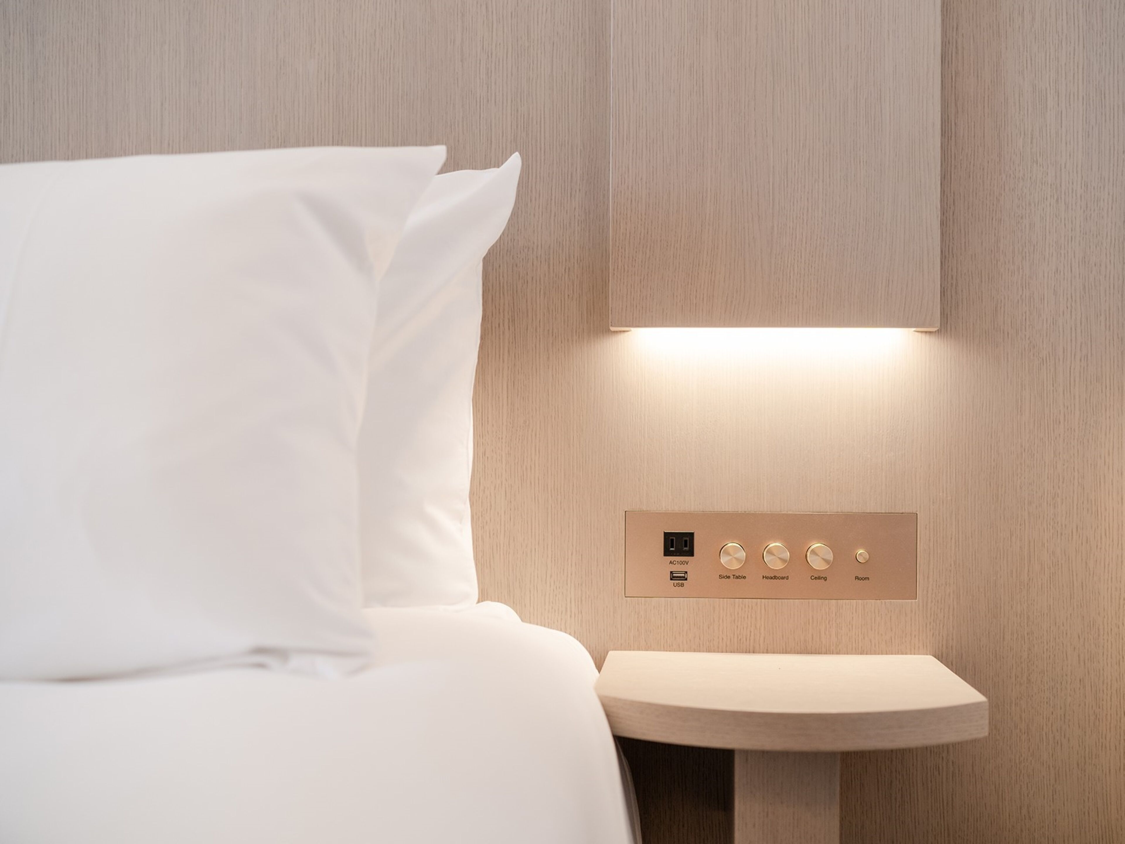 A modern hotel room with a bed, a bedside table, and a control panel with buttons for side table, headboard, ceiling, and room.