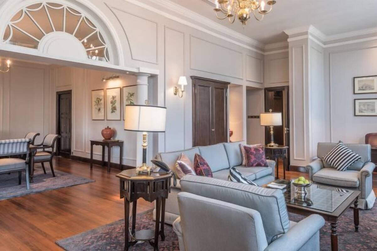A spacious living room with a large grey sofa, a coffee table, and a chandelier. There are two armchairs, a small side table, and a lamp. The room has wooden flooring and white walls with decorative molding.