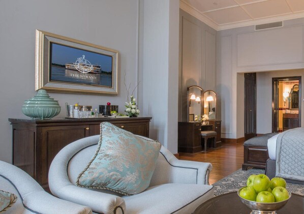 A hotel room with a large window, a framed picture of a ship, a dining table with a bowl of apples, and a bed with a quilt.