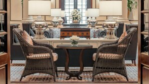 A formal sitting room with a round table, two wicker chairs, a central console table with lamps, and a large window with a view. - The Strand Yangon (Yangon)