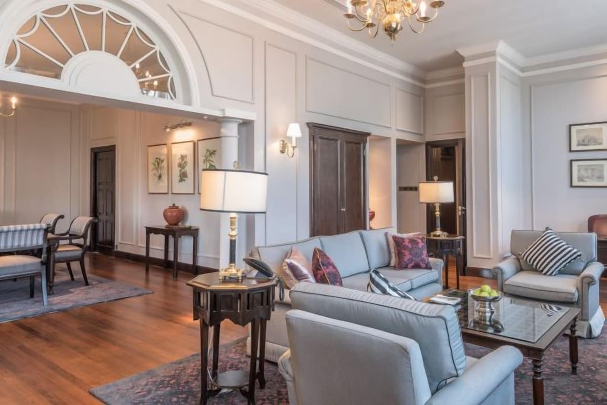 Un salón espacioso con un sofá grande de color gris, una mesa de centro y una araña. Hay dos sillones, una mesita auxiliar y una lámpara. El suelo es de madera y las paredes blancas con molduras decorativas.