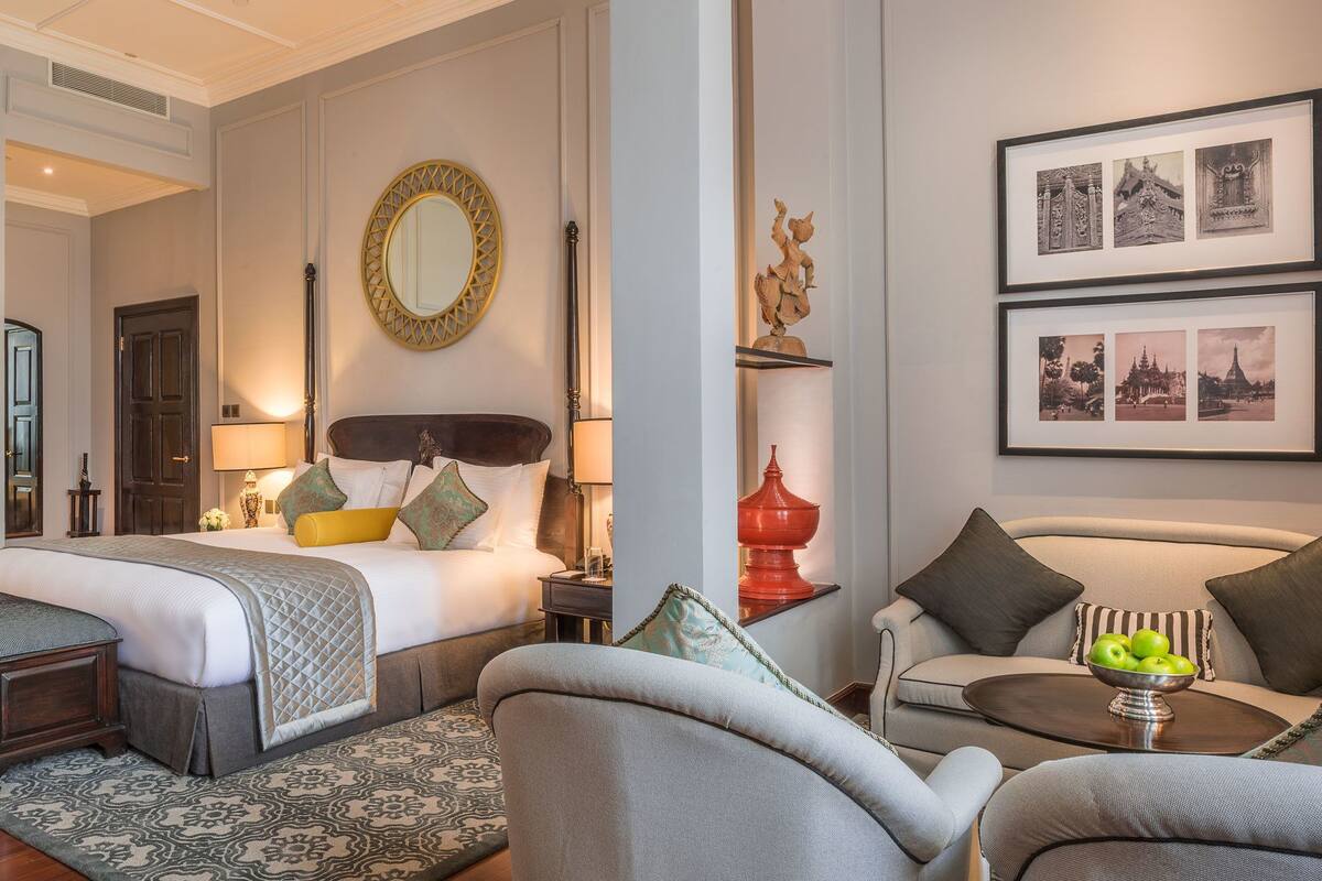 A hotel room with a large bed, a sofa, a coffee table with a bowl of fruit, and a wall with framed pictures.