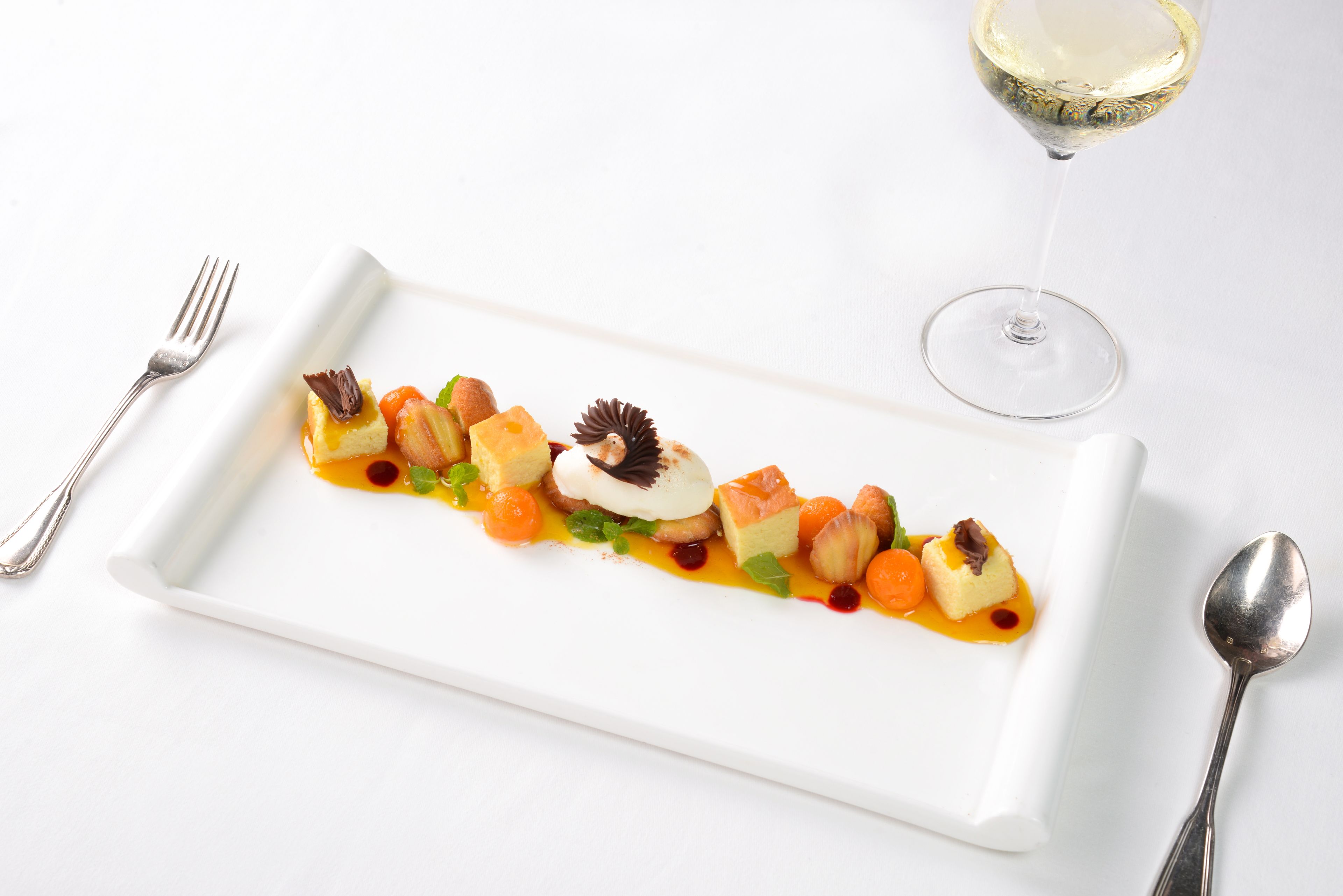 Un plato rectangular blanco con un postre, acompañado de un tenedor, una cuchara y una copa de vino sobre una superficie blanca.