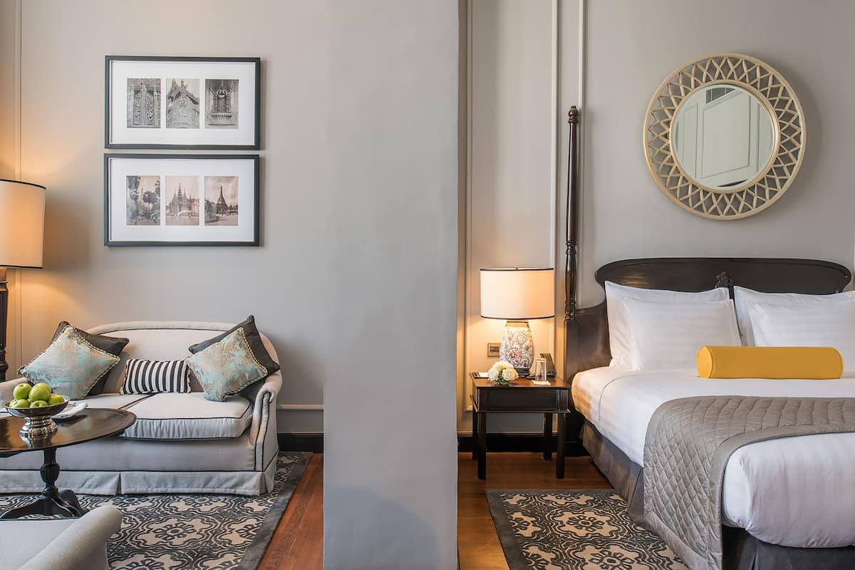 A hotel room with a bed, bedside tables, a sofa, a round table with fruit, and framed pictures on the wall.