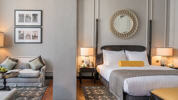A hotel room with a bed, bedside tables, a sofa, a round table with fruit, and framed pictures on the wall.