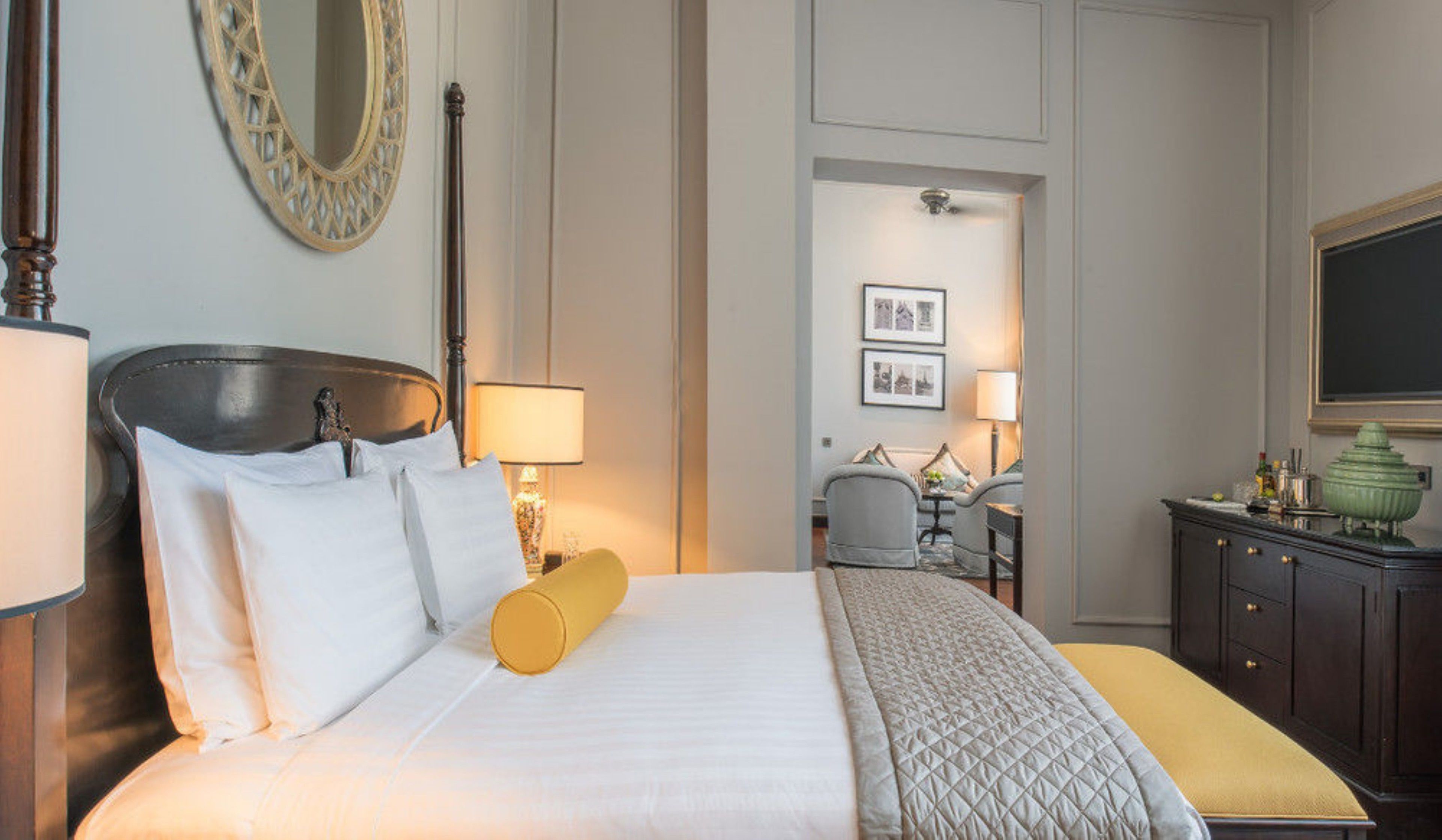 A neatly made bed with white linens and a yellow decorative pillow, a bedside lamp, a mirror with a decorative frame, and a dresser with a green vase.