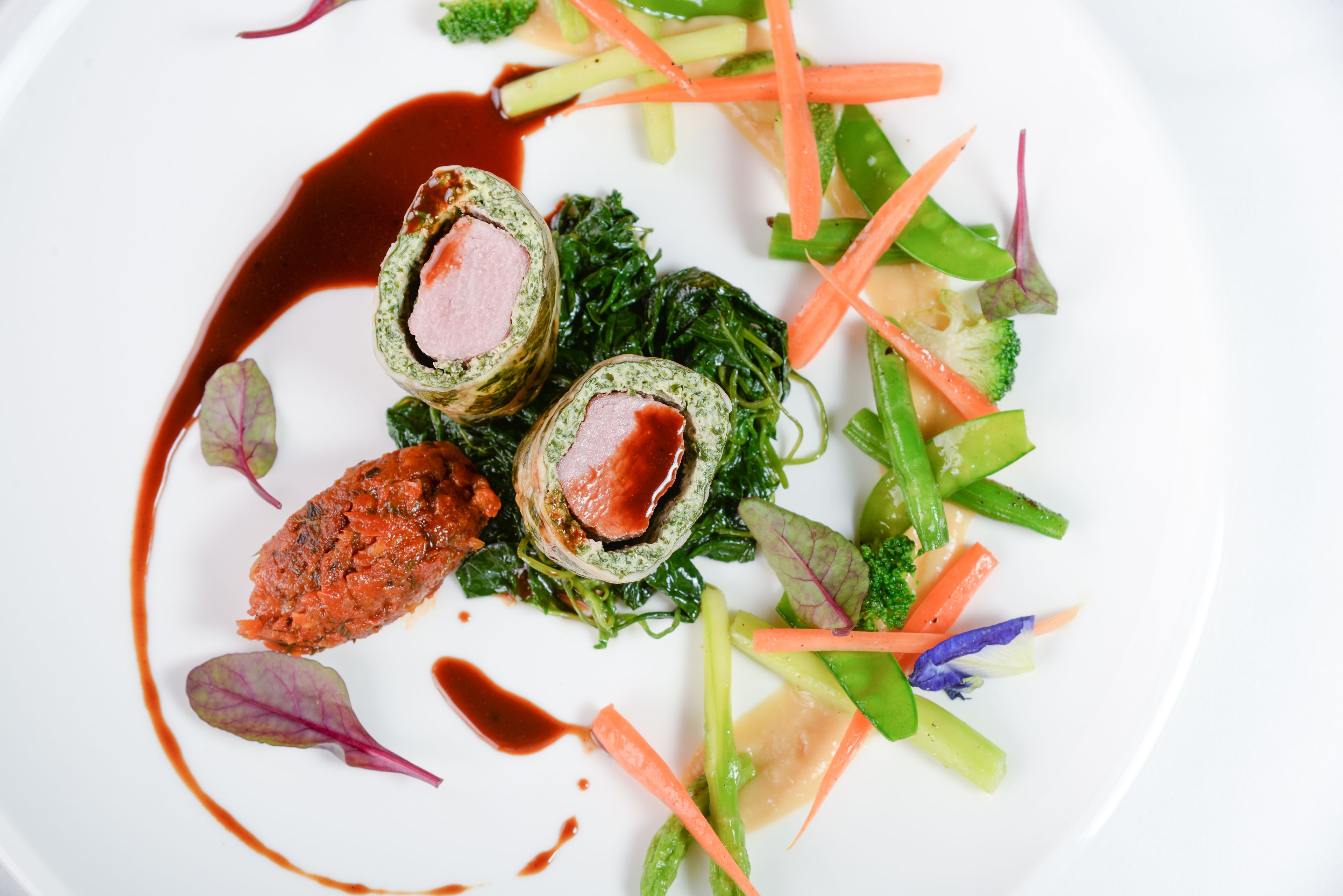 A white plate with a dish consisting of rolled meat, greens, and vegetables, with a dark sauce.