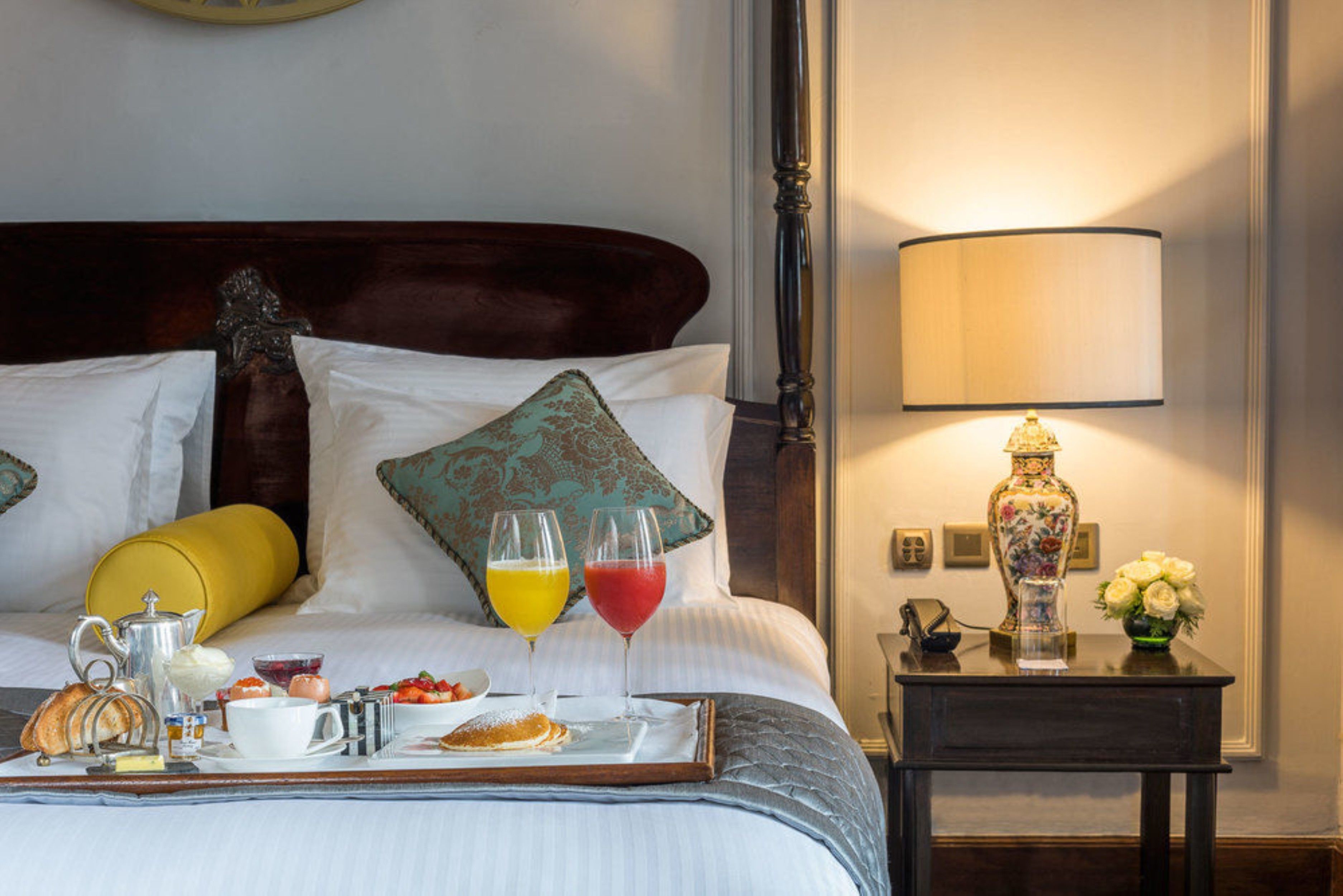 Een net opgemaakt bed met kussens, een nachtkastje met een lamp en een dienblad met ontbijtartikelen.