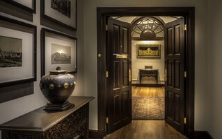Un pasillo con puertas de madera oscura, un gran jarrón decorativo sobre una consola y cuadros en las paredes.