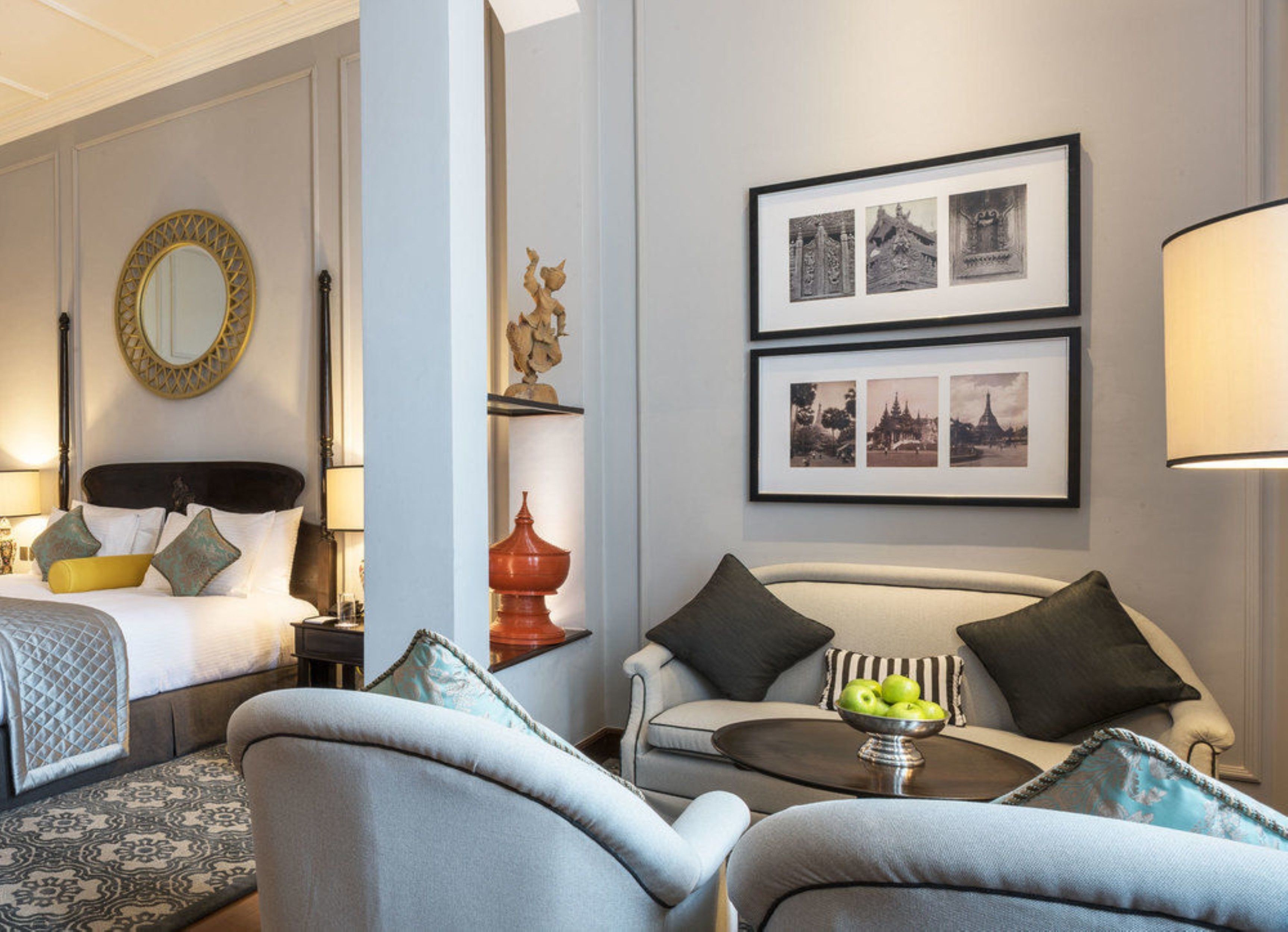 A hotel room with a bed, a sofa, a coffee table with apples, a mirror, and framed pictures on the wall.
