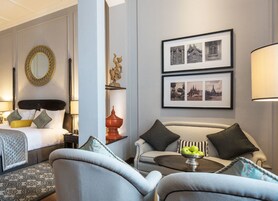 A hotel room with a bed, a sofa, a coffee table with apples, a mirror, and framed pictures on the wall.