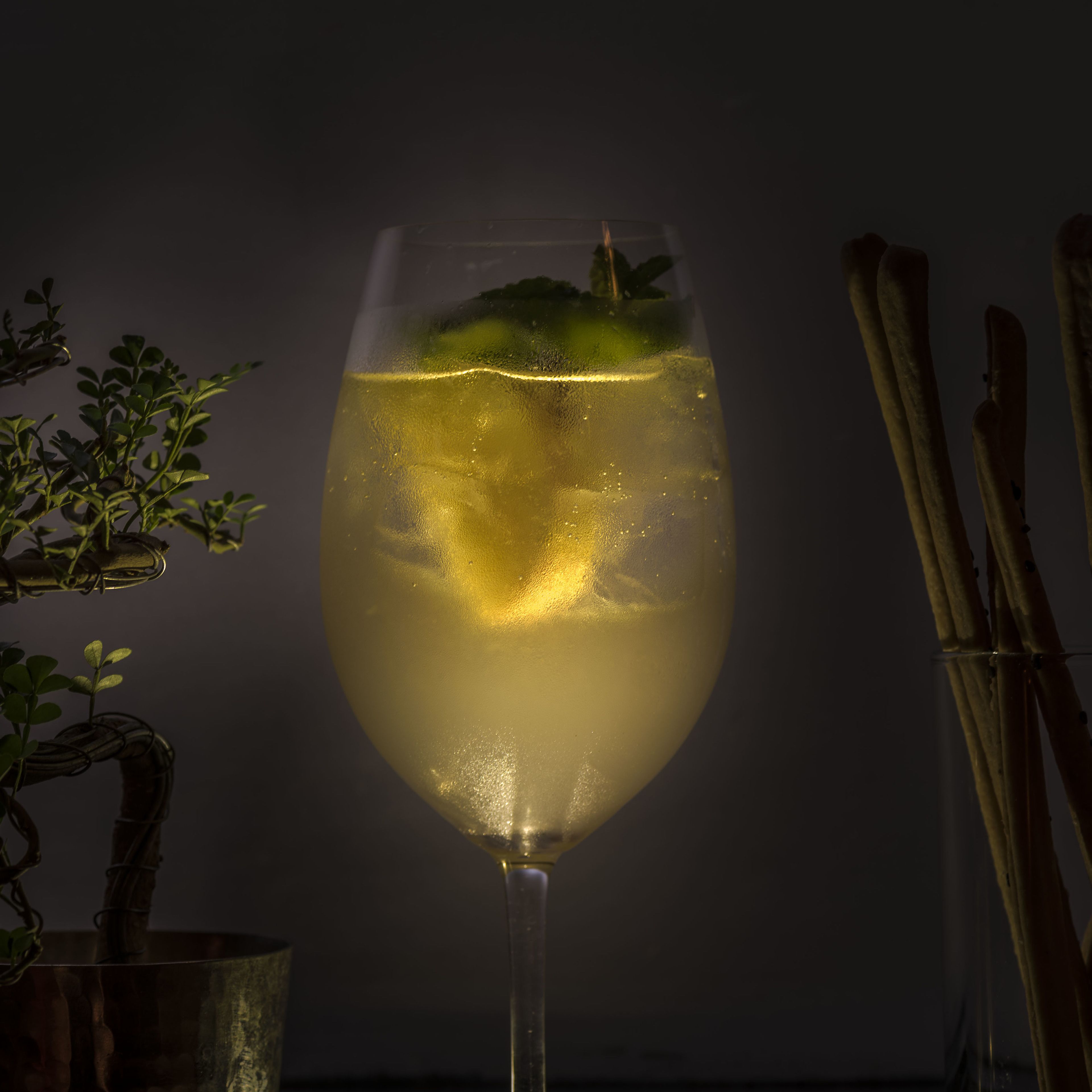 A glass of a clear, yellowish beverage with a garnish, possibly mint, in a stemmed glass.