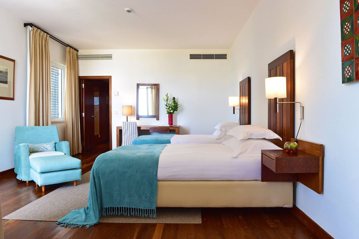 A hotel room with a bed, a chair, a desk, a mirror, and a wall-mounted lamp.