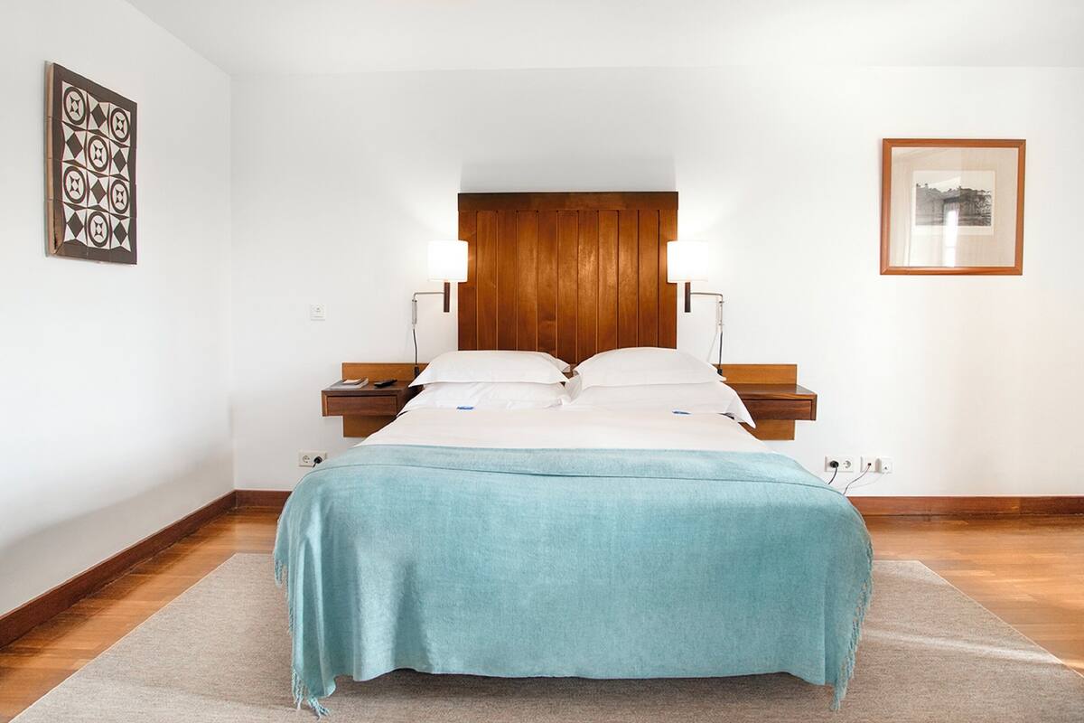 A neatly arranged bedroom with a wooden headboard, a bed with a teal bedspread, bedside tables with lamps, and a framed picture on the wall.