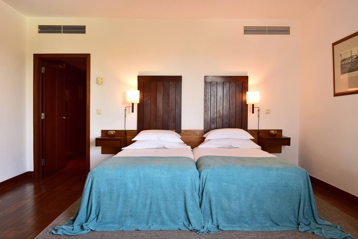 A double bed with a teal bedspread, two bedside tables with lamps, a wooden door, and a framed picture on the wall.