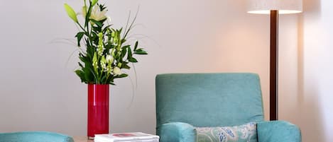 A room with teal upholstered chairs, a red vase with flowers, a geometric wall art piece, and a floor lamp.