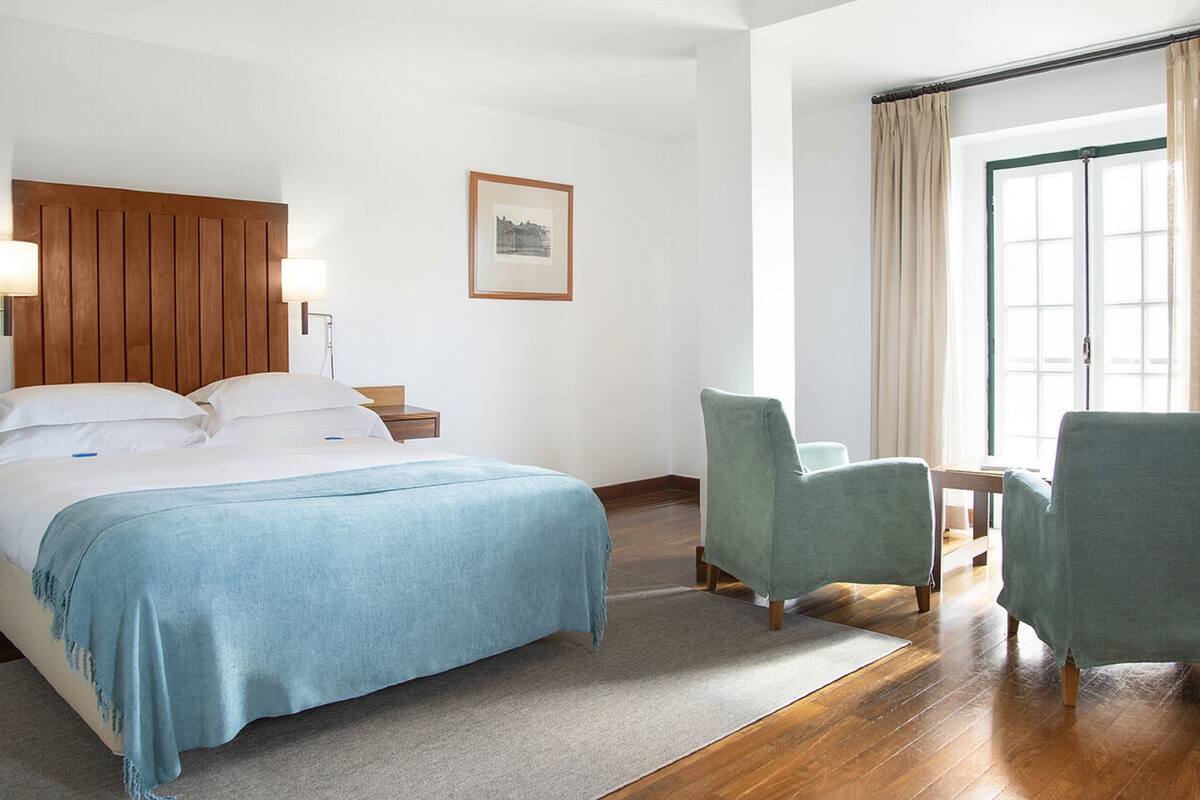 A hotel room with a bed, two chairs, a small table, a nightstand, a lamp, a framed picture, and a window with curtains.