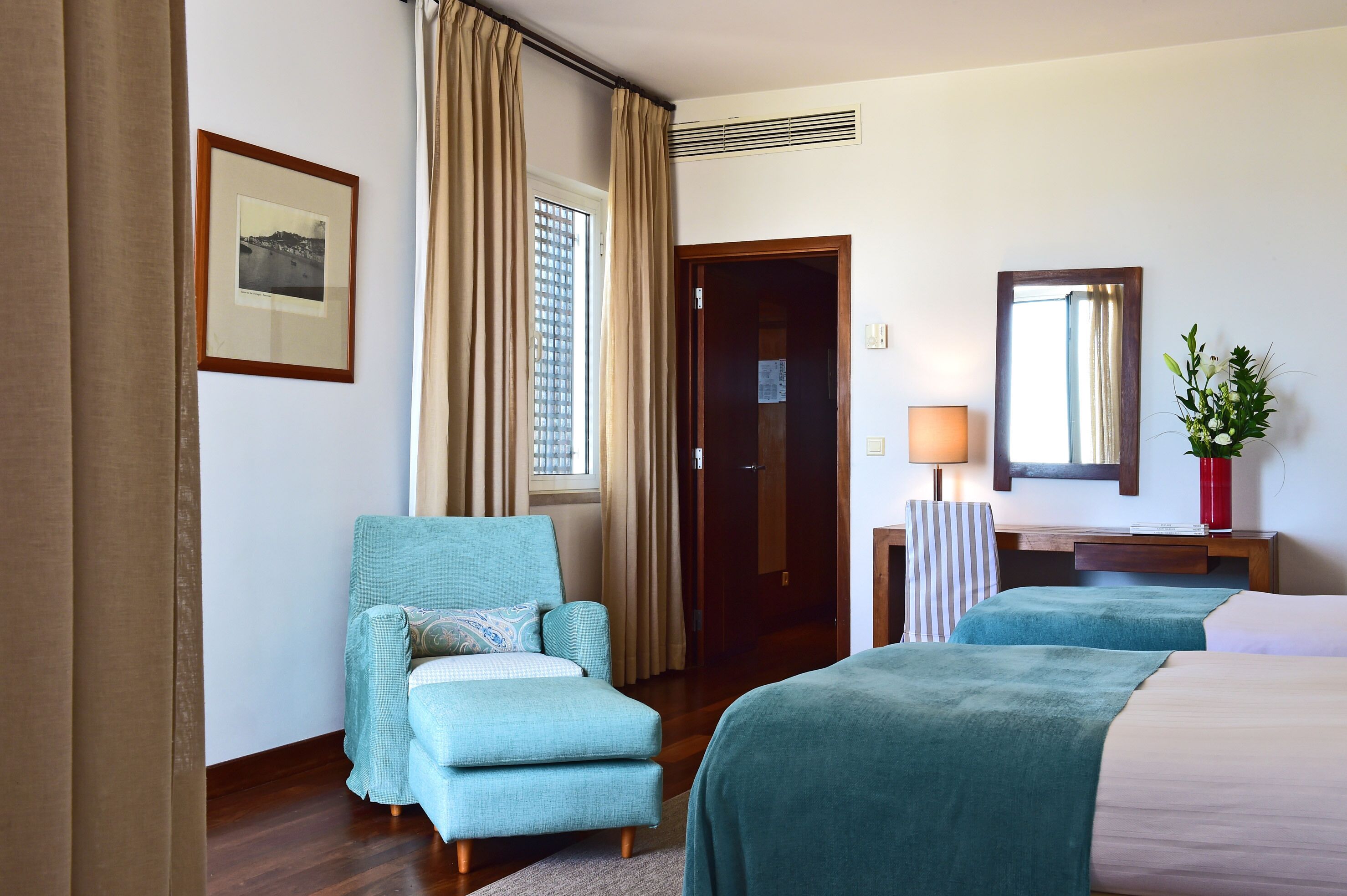 A hotel room with a bed, a chair, a desk, a lamp, a mirror, and a window with curtains.