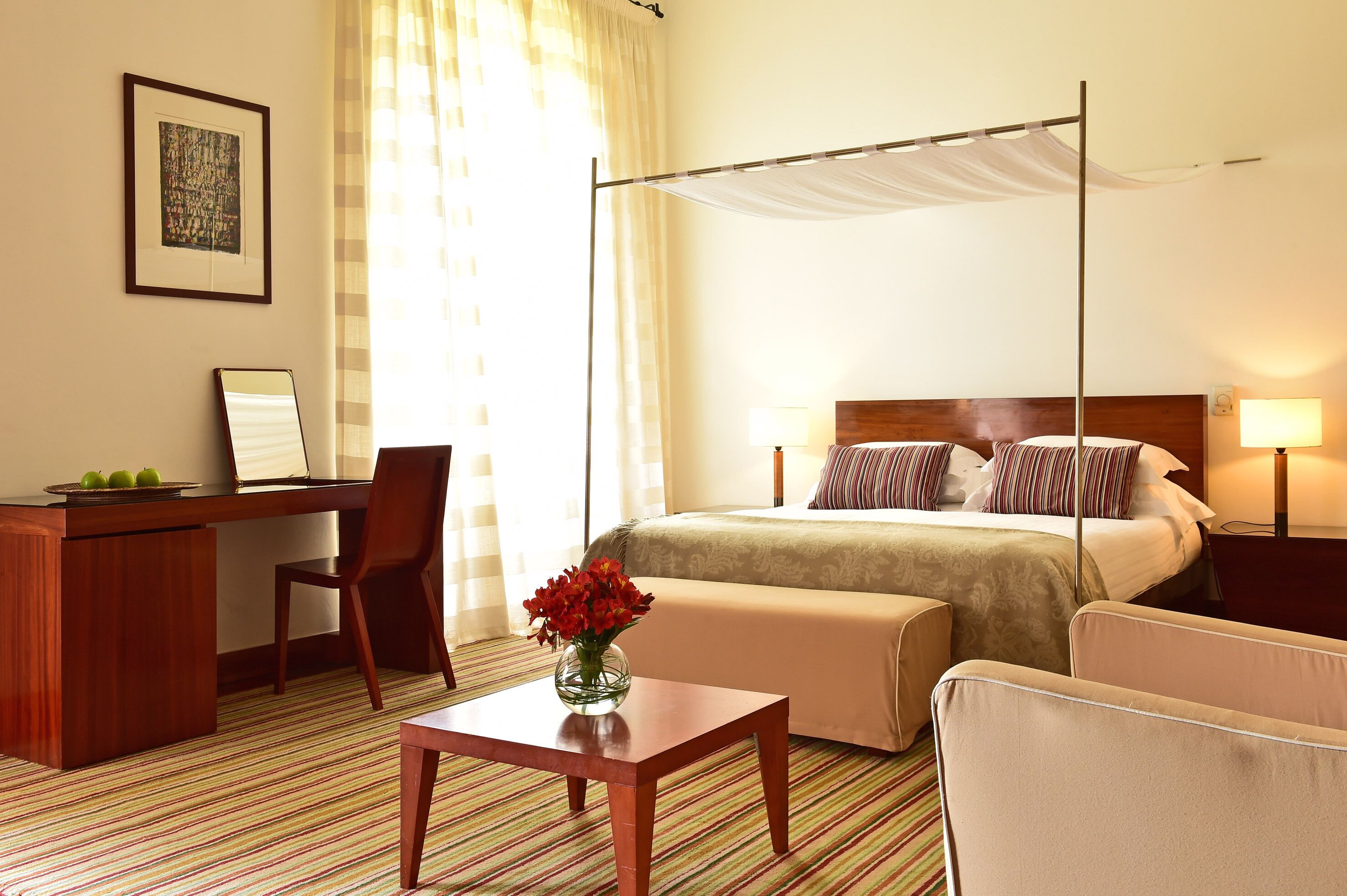 A hotel room with a bed, a desk, a chair, a small table, and a vase of flowers.